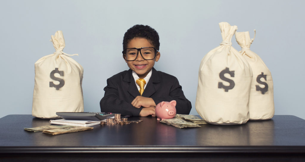 young boy in suit and tie sitting at desk with money bags and piggy bank