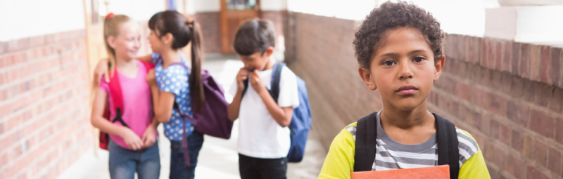 Male elementary student stands sadly in the hallway alone while classmates talk in the background.