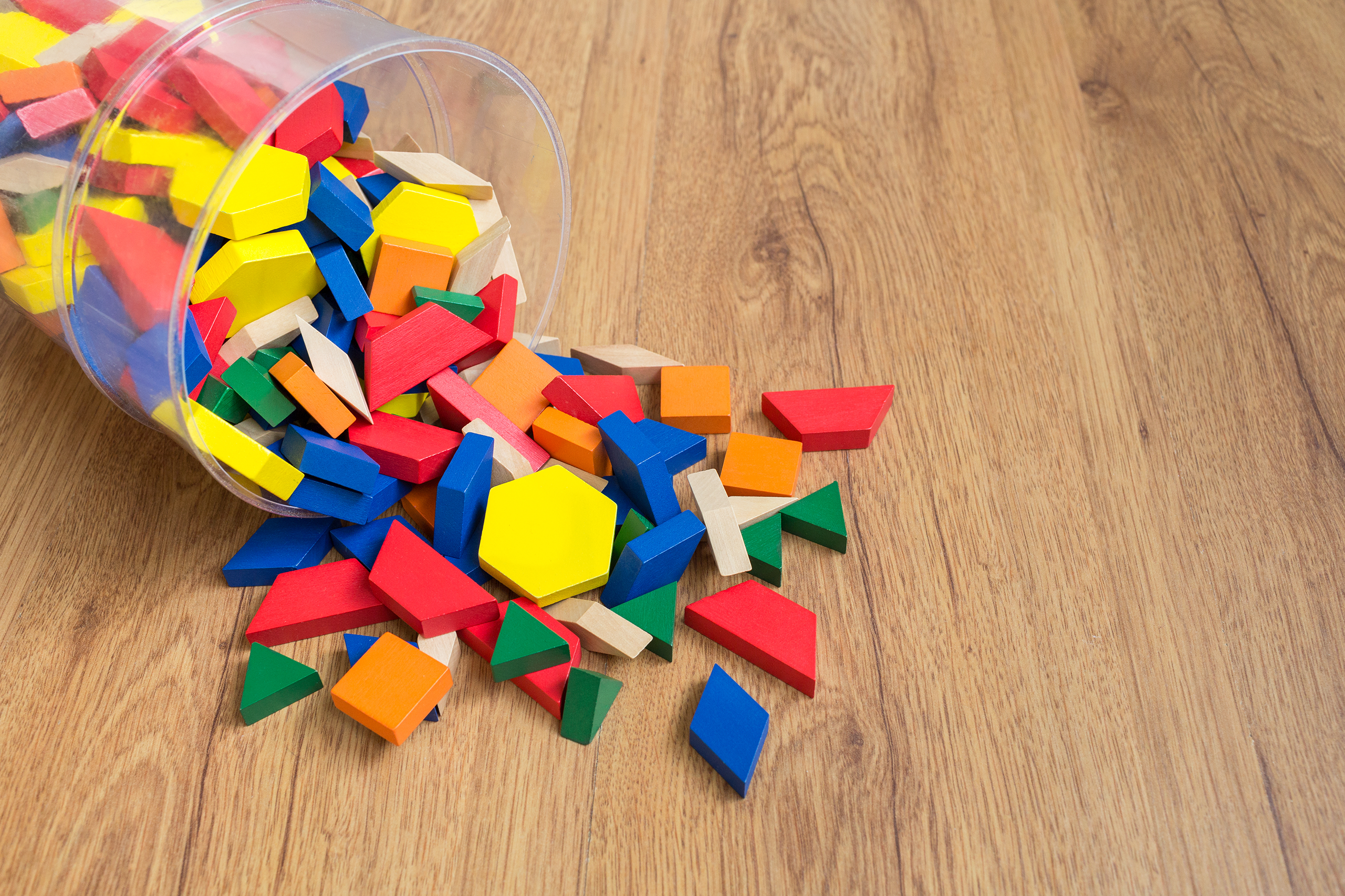 A clear container full of colorful pattern blocks spilled on a wooden surface.