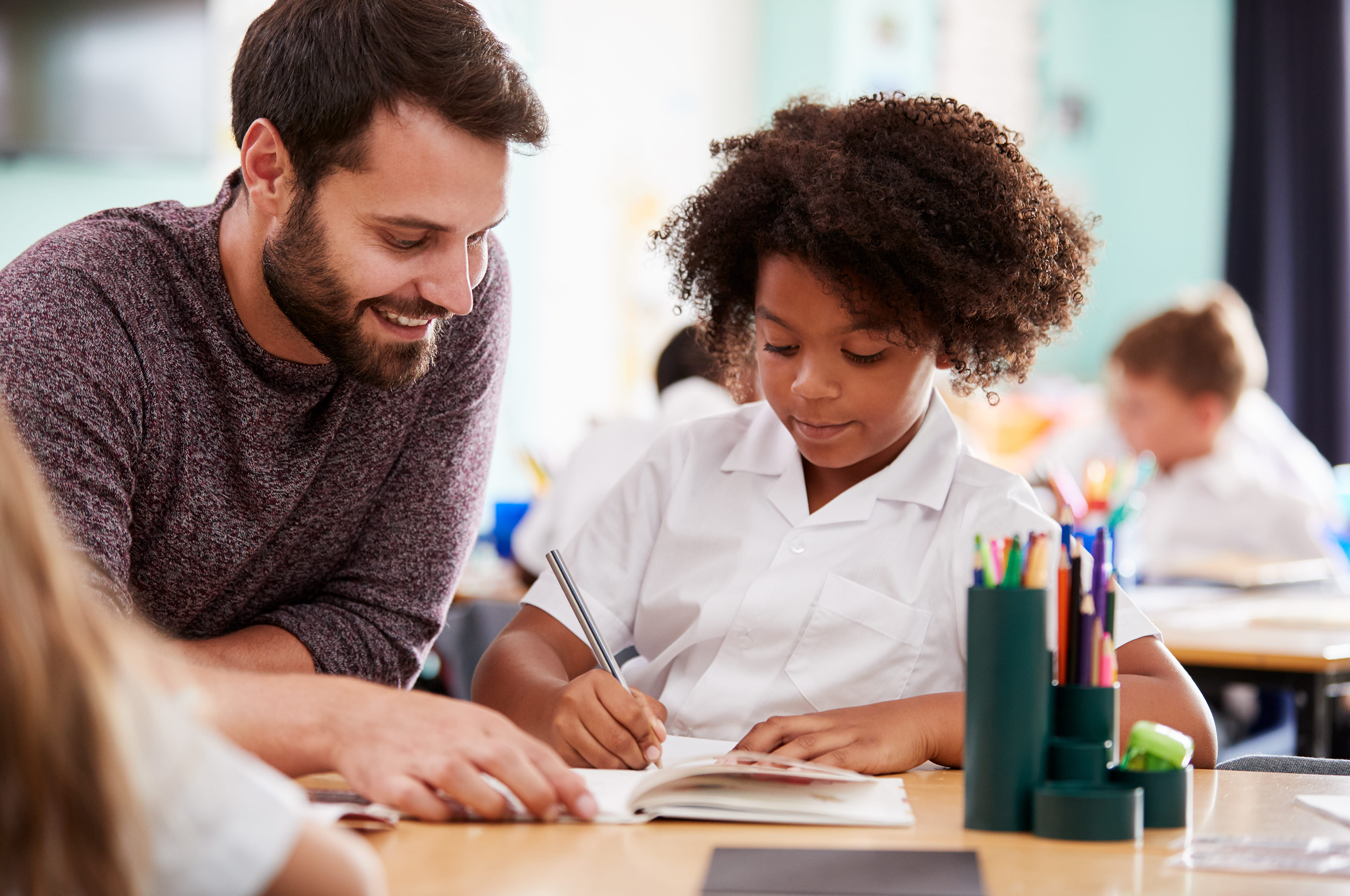 Male elementary school teacher supports student as they write in a journal.