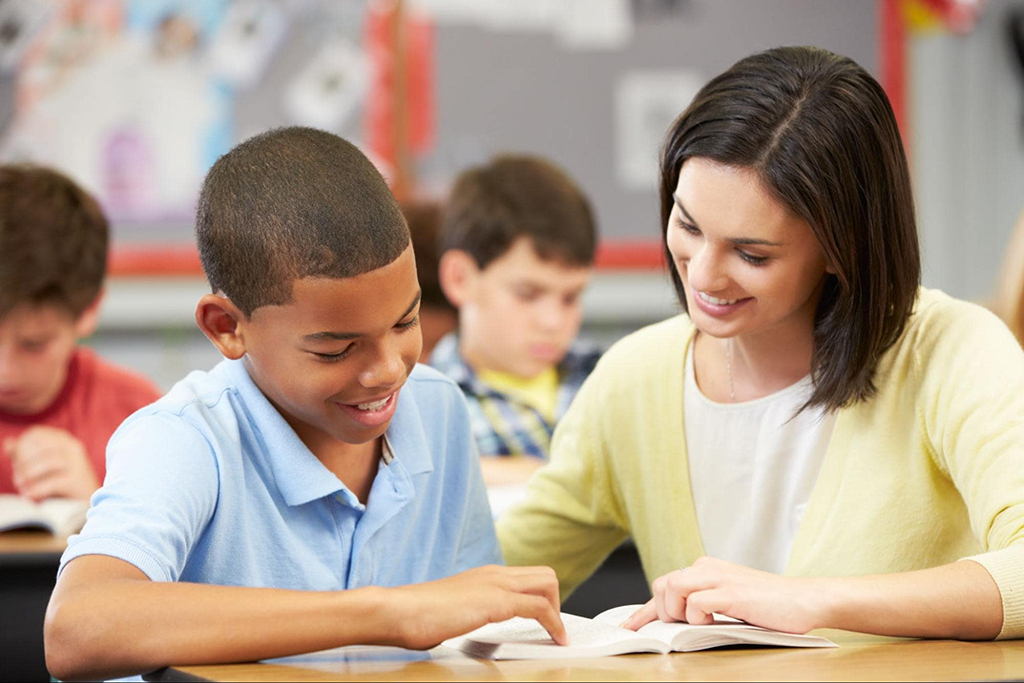 Female teacher assists male student as he reads aloud.