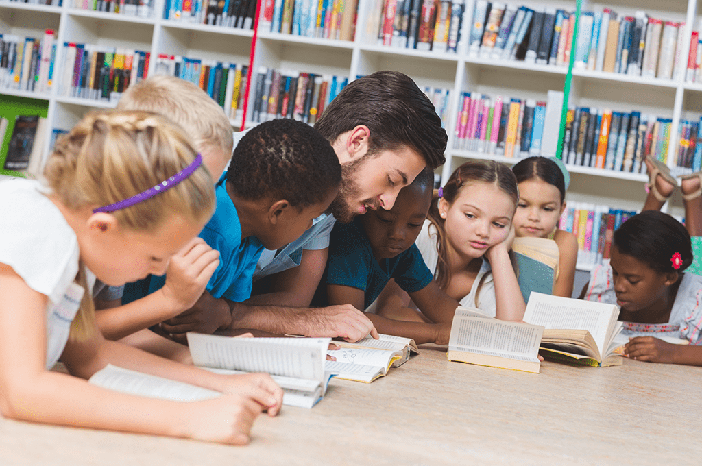 Male teacher reads aloud to group of students at a library table.