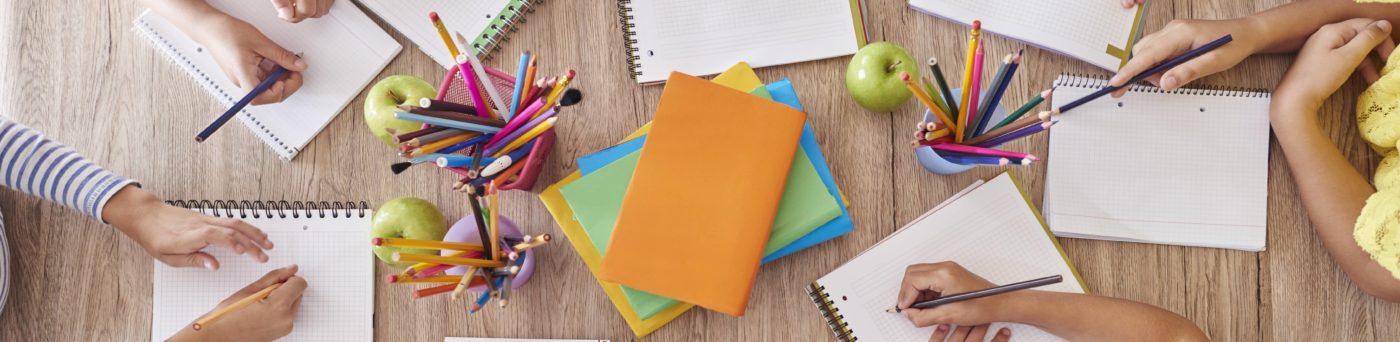 An aerial shot of students writing in notebooks at a table, with school supplies on it