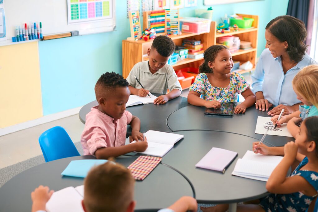 Group of elementary students working at a table in class. A little girl looks up at her teacher and smiles.