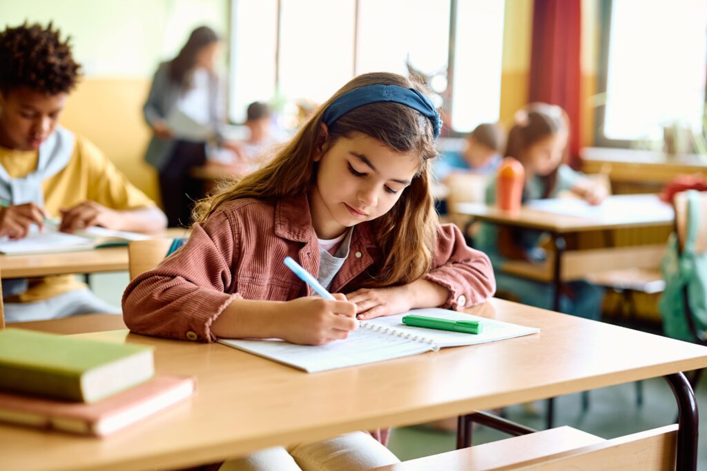 Young girl writing in a notebook at her desk in class.