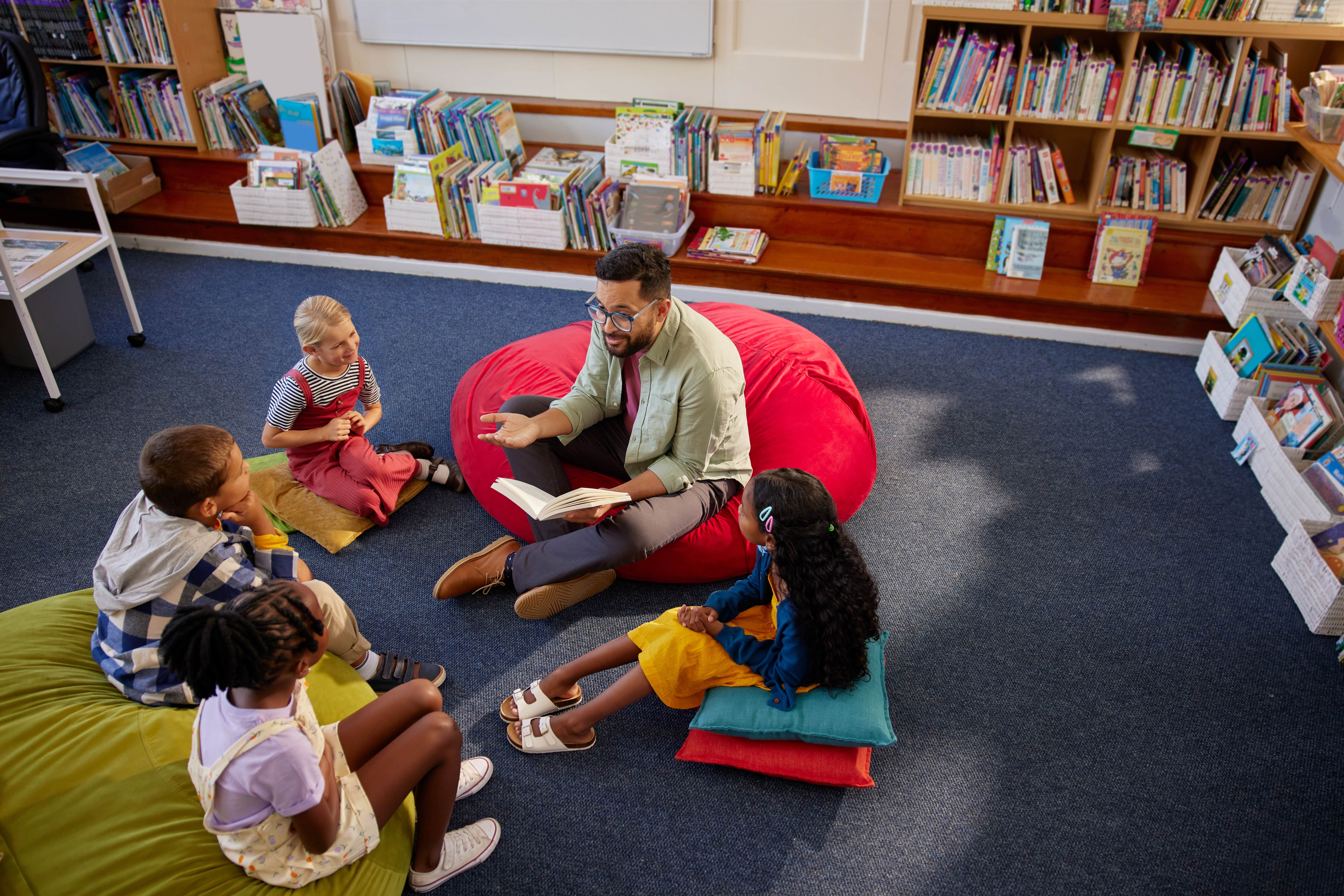 Teacher sitting on the floor in a bean bag chair reading to a group of students during story hour.