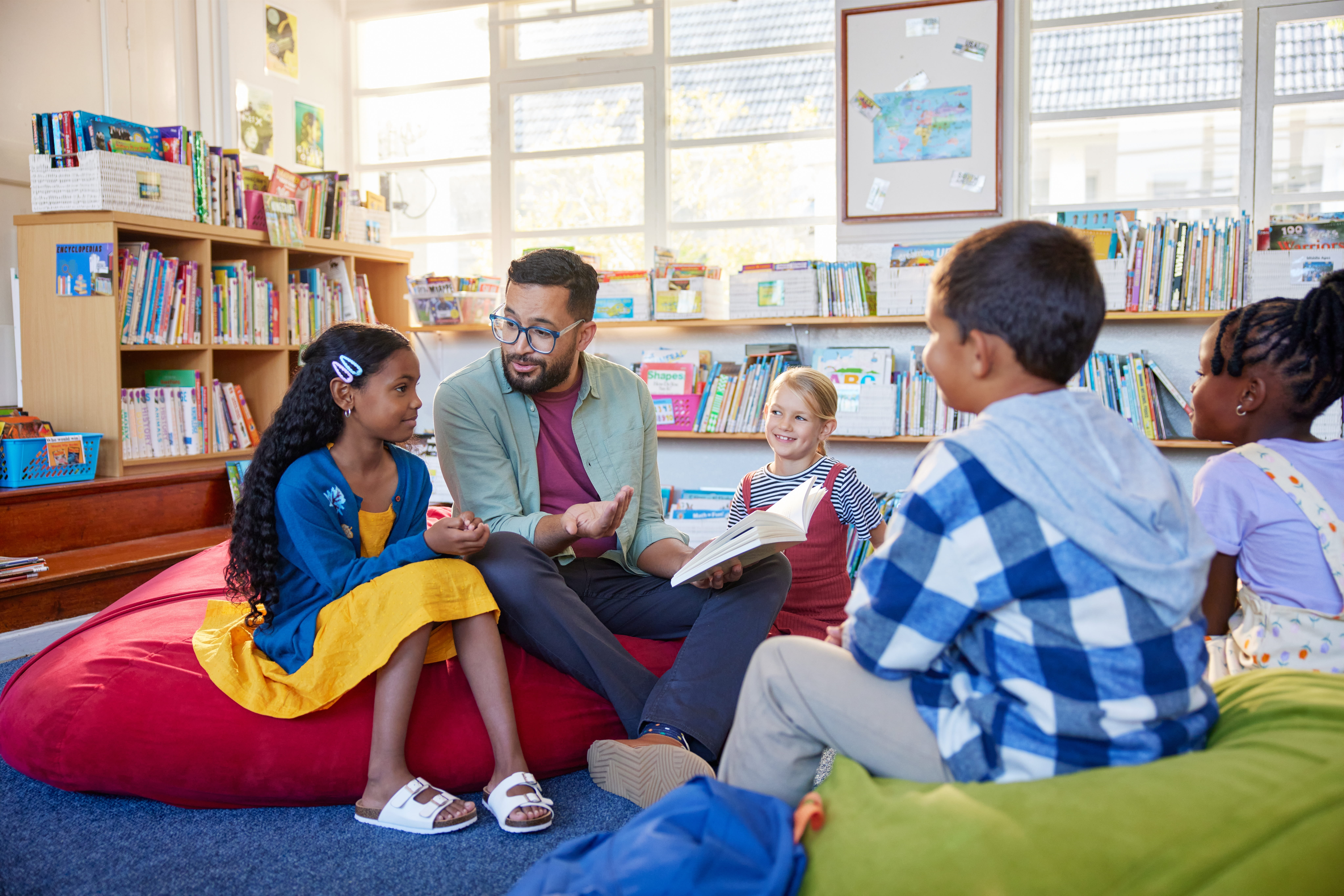 male teacher reading to a group of young students sitting in beanbag chairs in a library